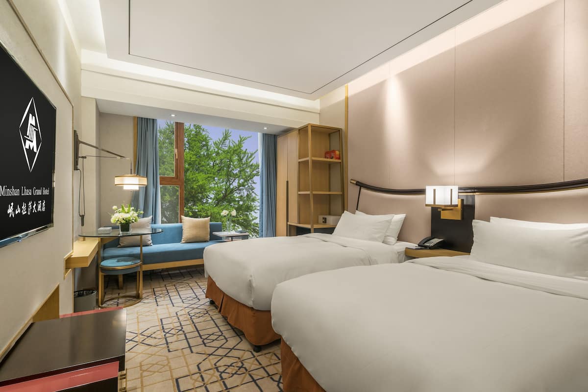 A hotel room with two beds, a sofa, a small table with a lamp, a TV, and a window with a view of trees.