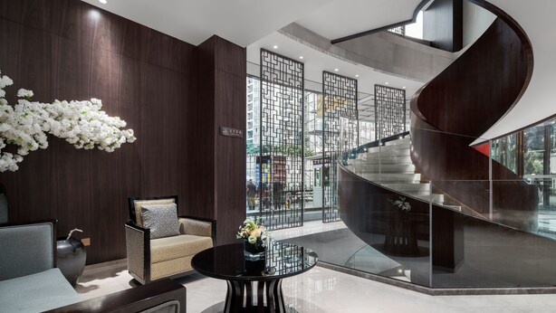 A modern lobby with a curved staircase, a seating area with a vase of flowers, and a view through a glass partition. - Minshan Lhasa Grand Hotel (Chengdu)