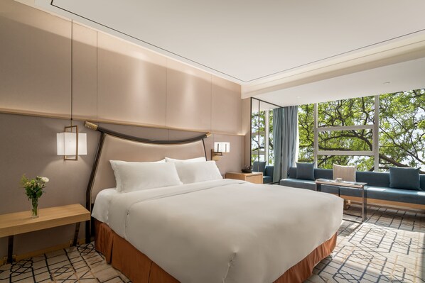 A modern hotel room with a large bed, a seating area with a sofa and armchairs, a small table, and a view of trees through large windows.