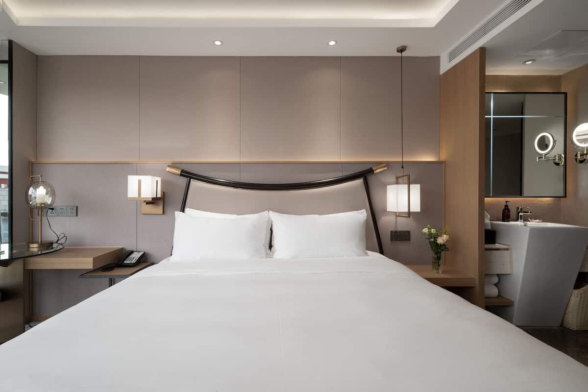 A modern hotel room with a large bed, bedside lamps, a nightstand with a phone, a mirror with lights, and a bathroom with a sink and mirror.