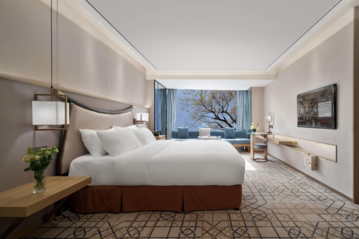A modern hotel room with a large bed, a desk, a chair, and a view of the outdoors.