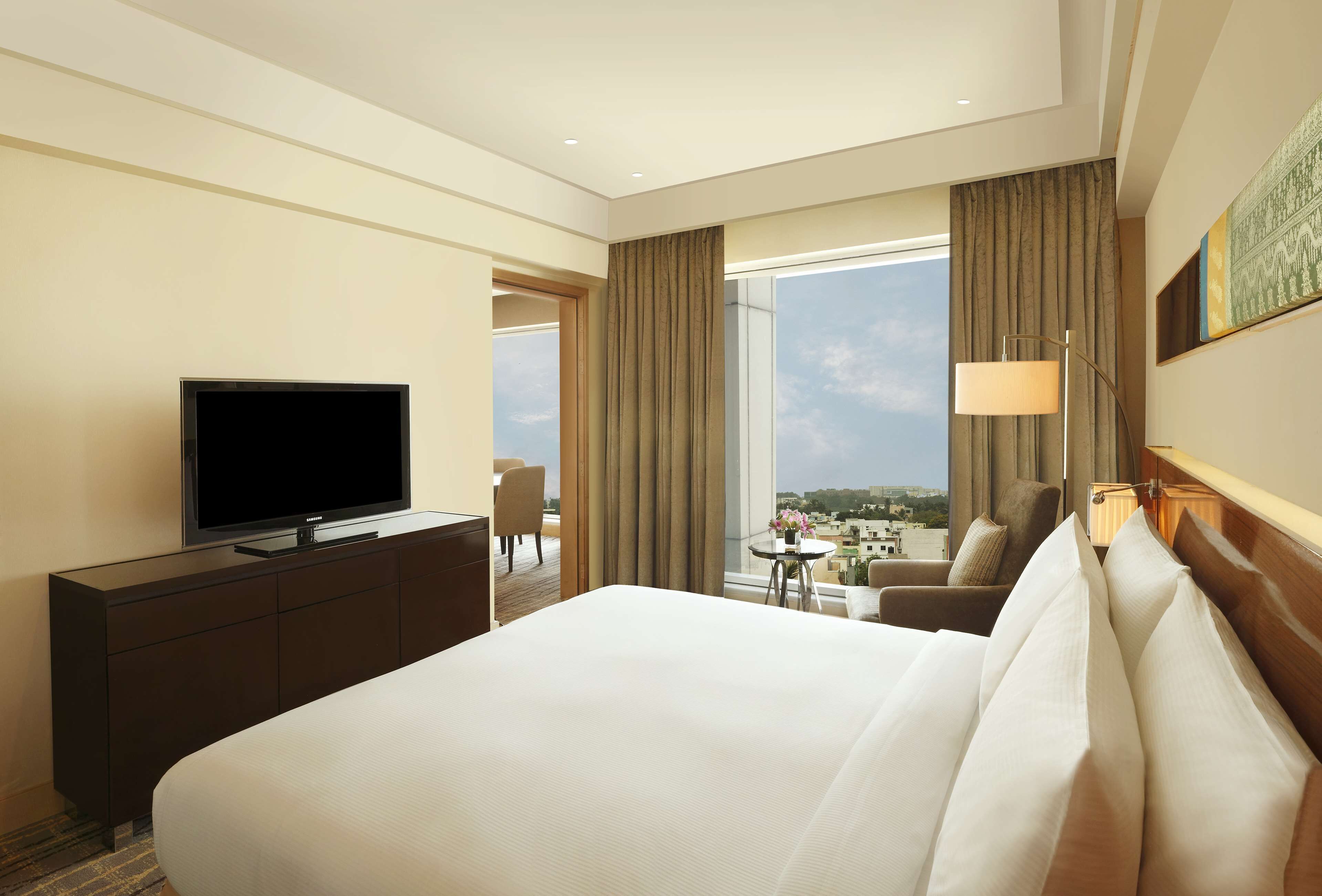 A hotel room with a large bed, a television on a dresser, a desk with a lamp, and a view of the city through large windows.
