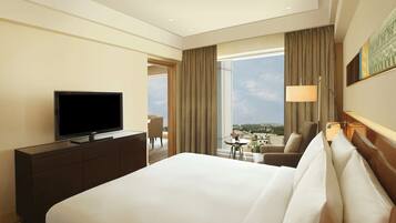 A hotel room with a large bed, a television on a dresser, a desk with a lamp, and a view of the city through large windows.