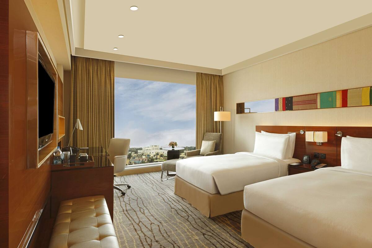Habitación de hotel con dos camas, televisión, escritorio con lámpara y vistas a la ciudad.