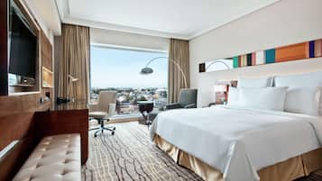 A hotel room with a large bed, a desk with a flat-screen TV, a chair, a bench, and a view of the city.