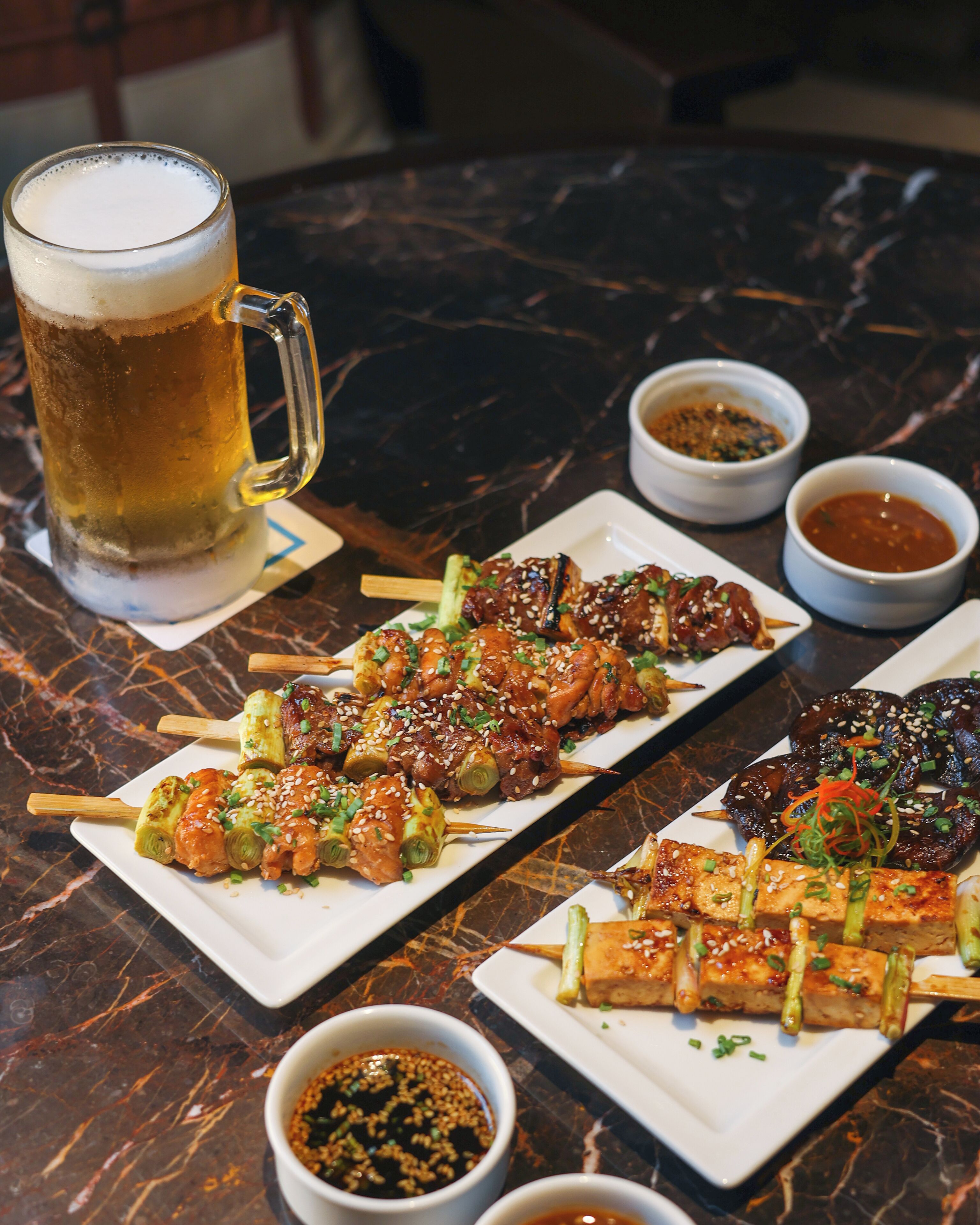 A table with skewers of food, a beer mug, and small bowls of sauces.