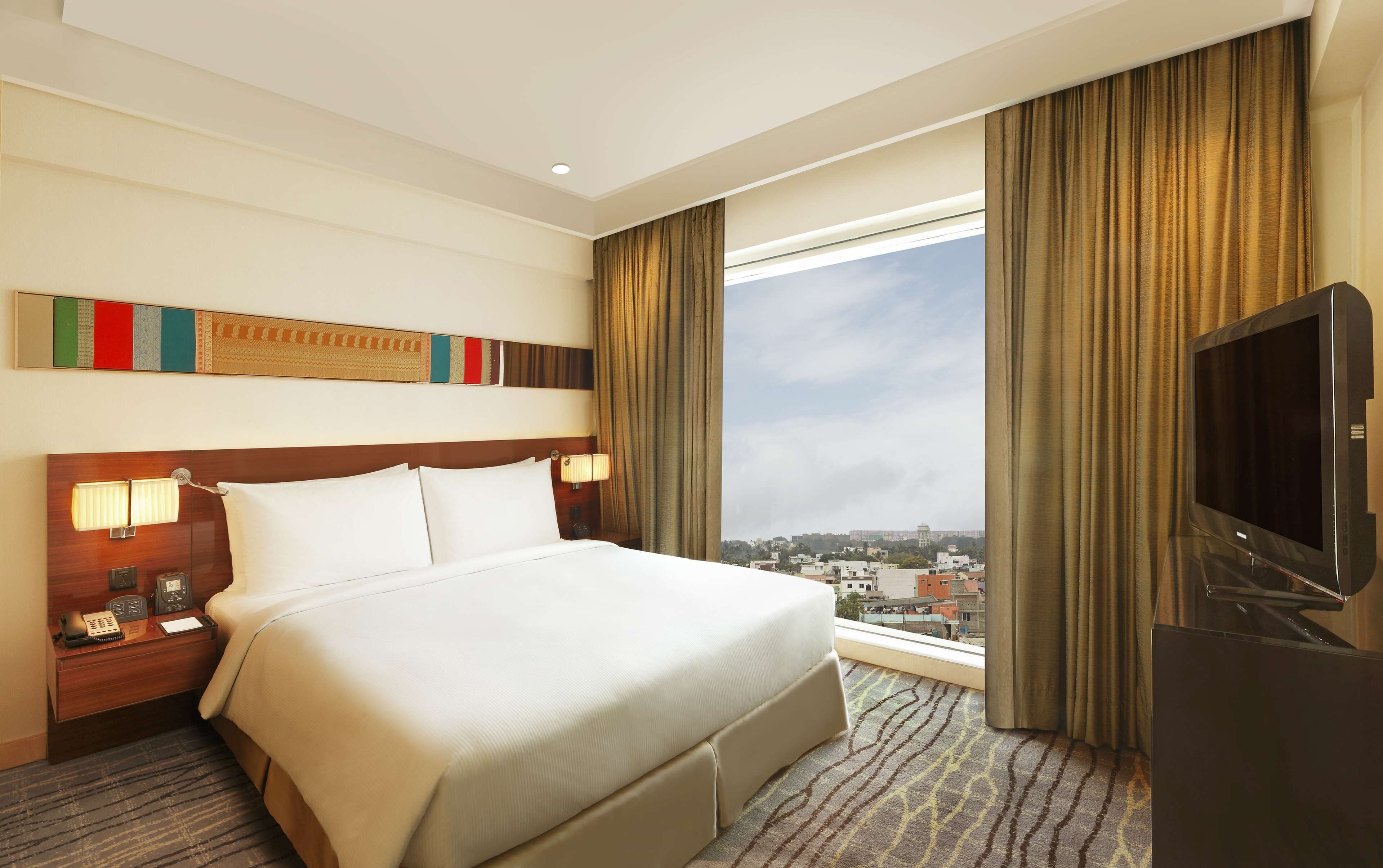 Ein Hotelzimmer mit einem großen Bett, einem Fernseher, einem Schreibtisch mit Telefon und einem Fenster mit Blick auf die Stadt.