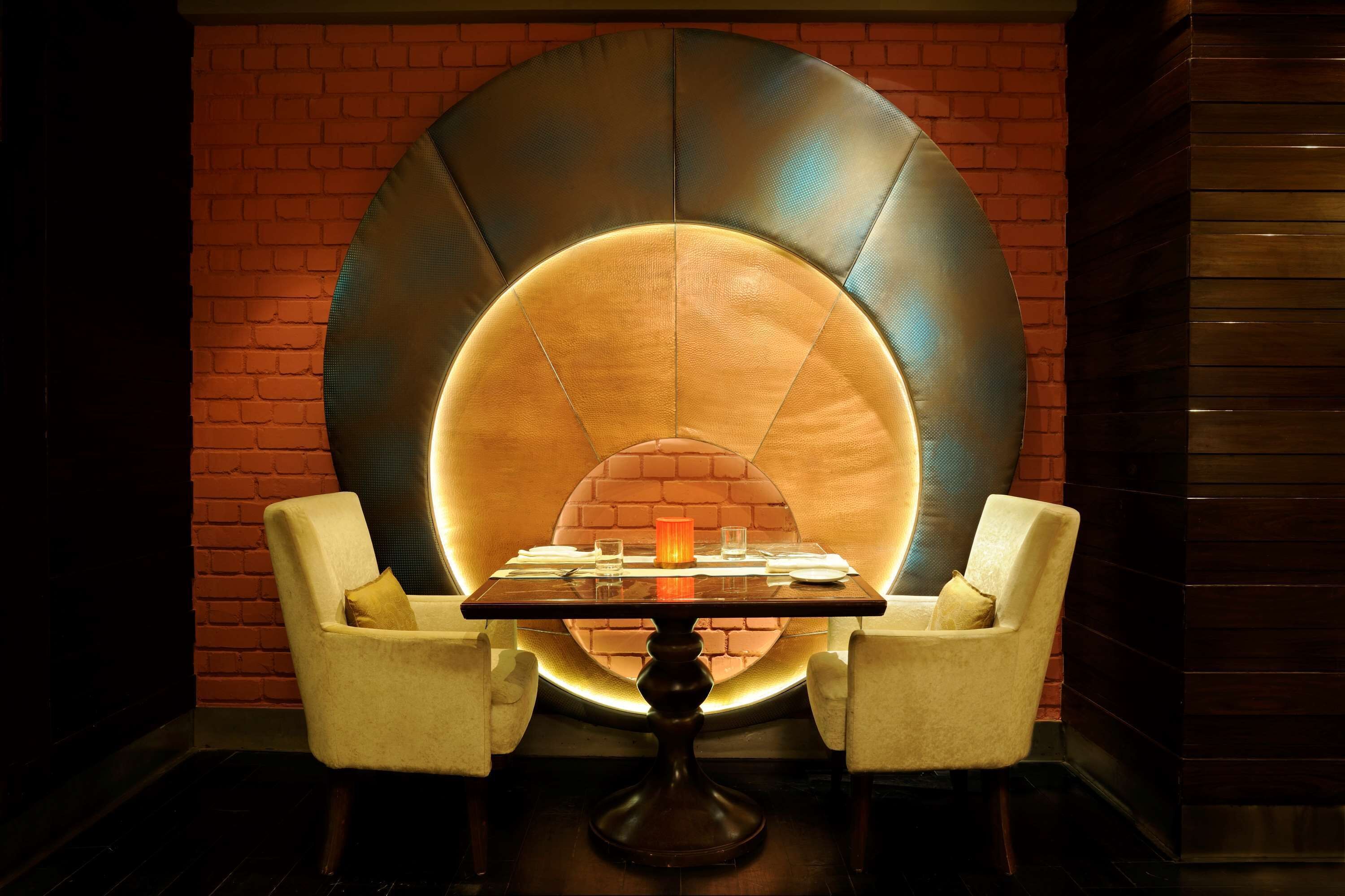 A dining area with a round table, two chairs, a candle, and a brick wall with a circular light fixture.