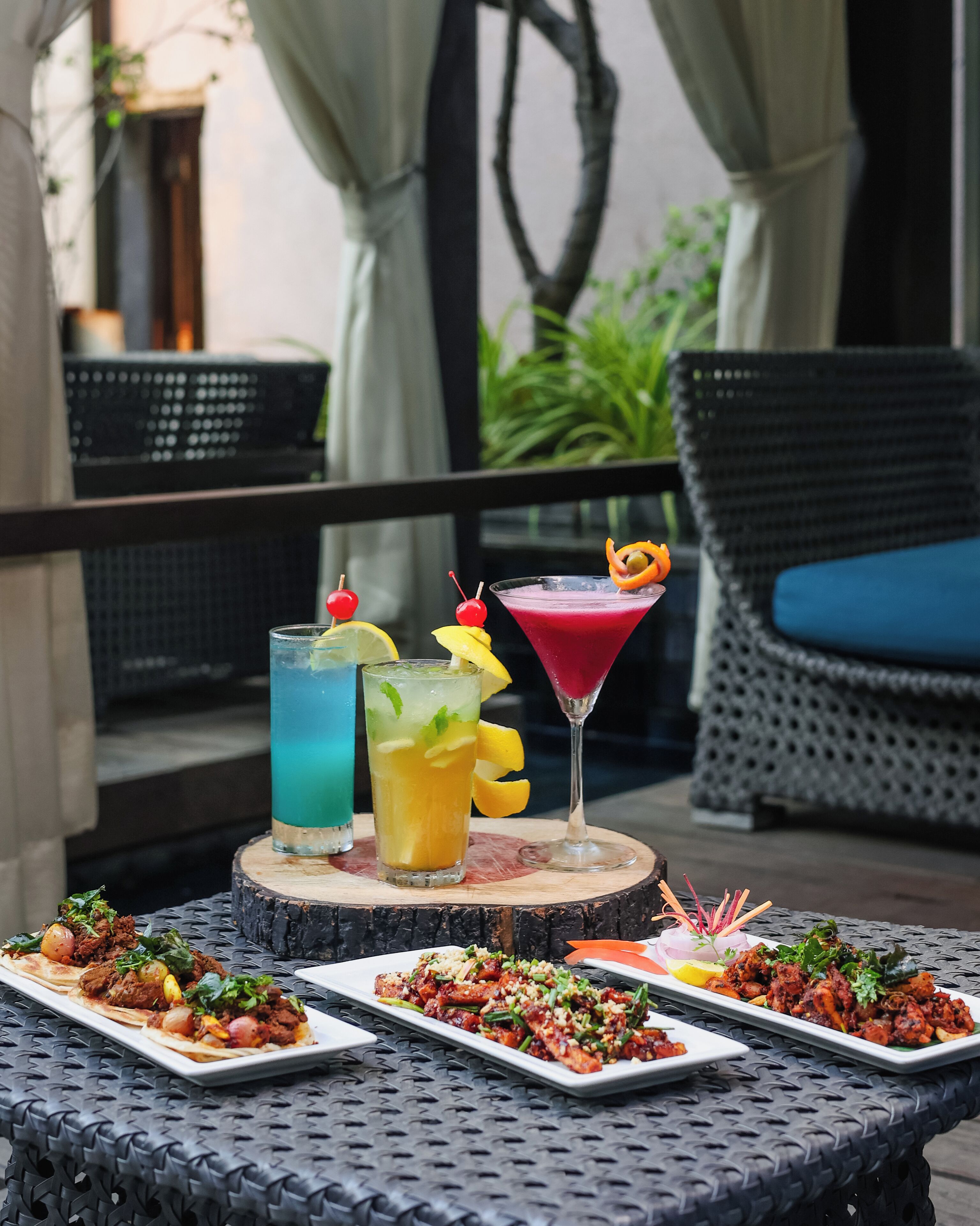 A table set with various dishes and drinks, a wicker chair, and a view of greenery in the background.