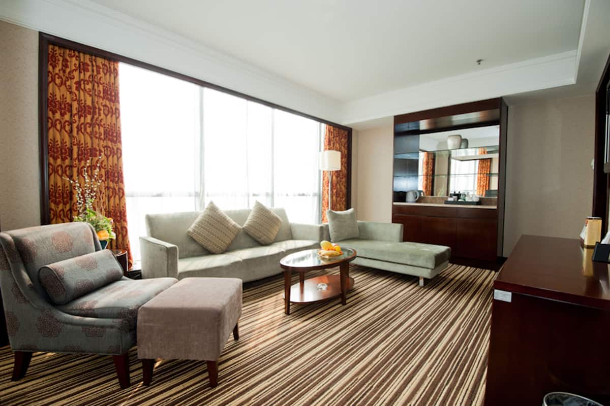 Quarto de hotel com sofá, poltrona e apoio para os pés, uma pequena mesa com abajur, uma janela grande com cortinas e uma cozinha visível através de um painel de vidro.