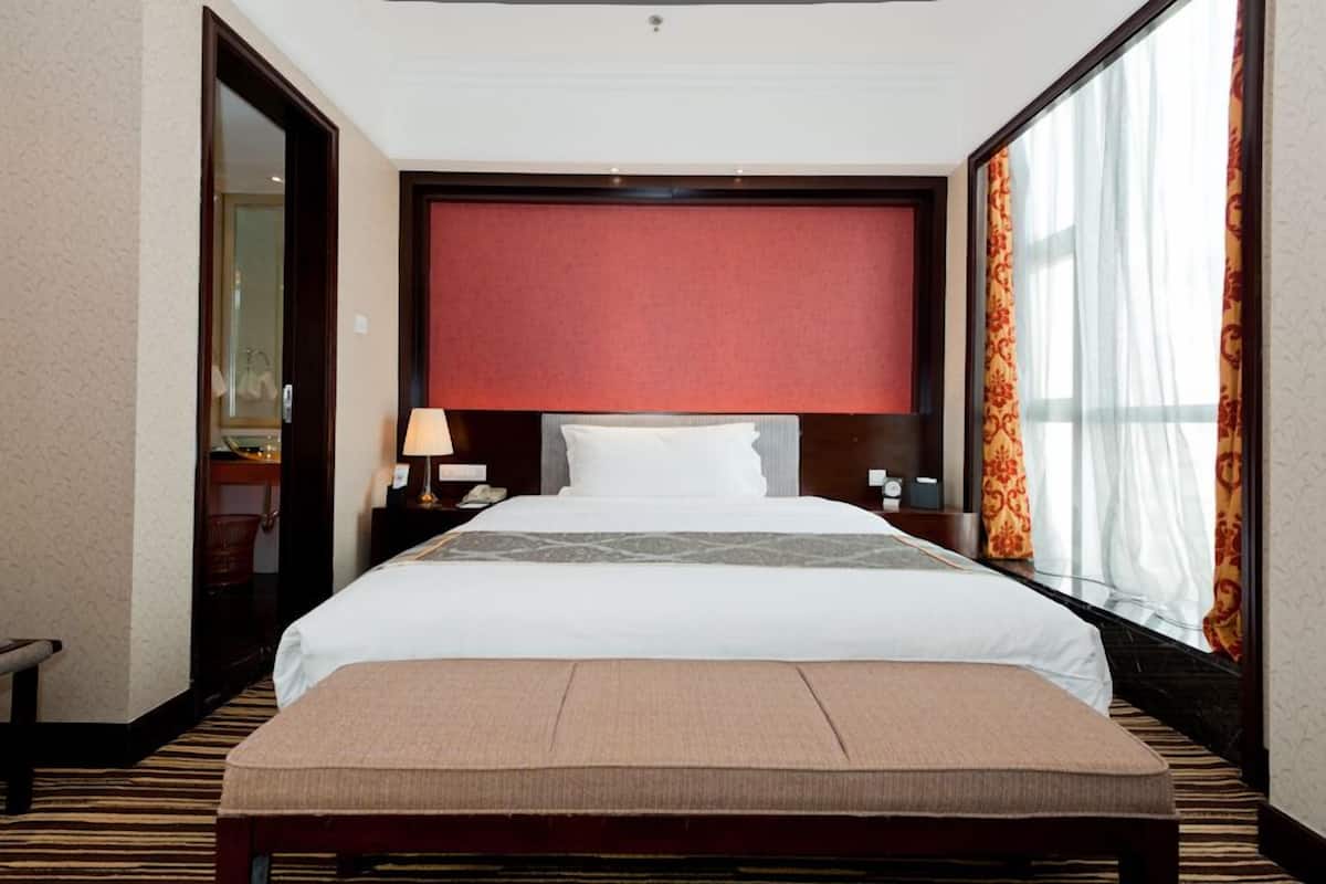 Quarto de hotel com uma cama grande, uma mesa de cabeceira com abajur, um banco no pé da cama e uma janela ampla com cortinas.