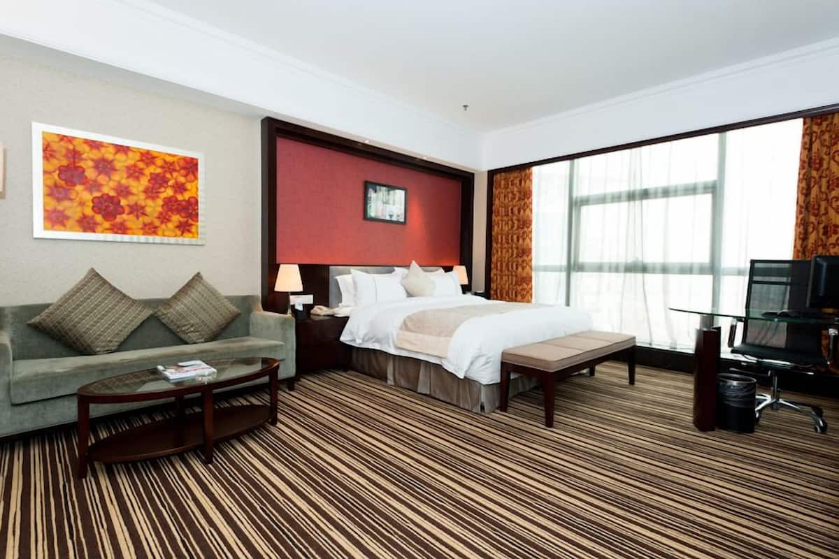 Quarto de hotel com cama, sofá, mesa de centro, escrivaninha e uma janela grande.