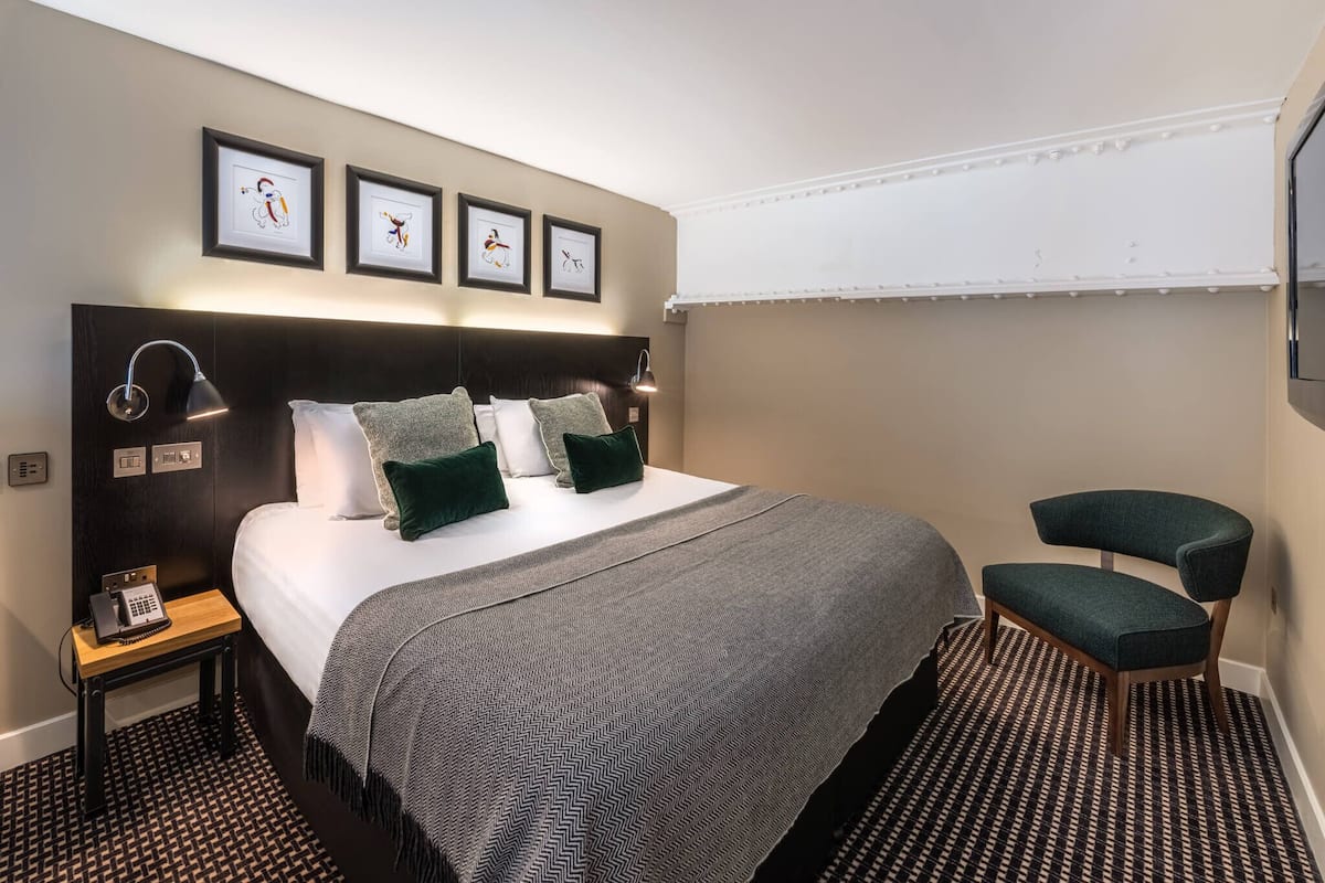 Ett välordnat hotellrum med en säng, sängbord, en stol och inramade konstverk på väggen.