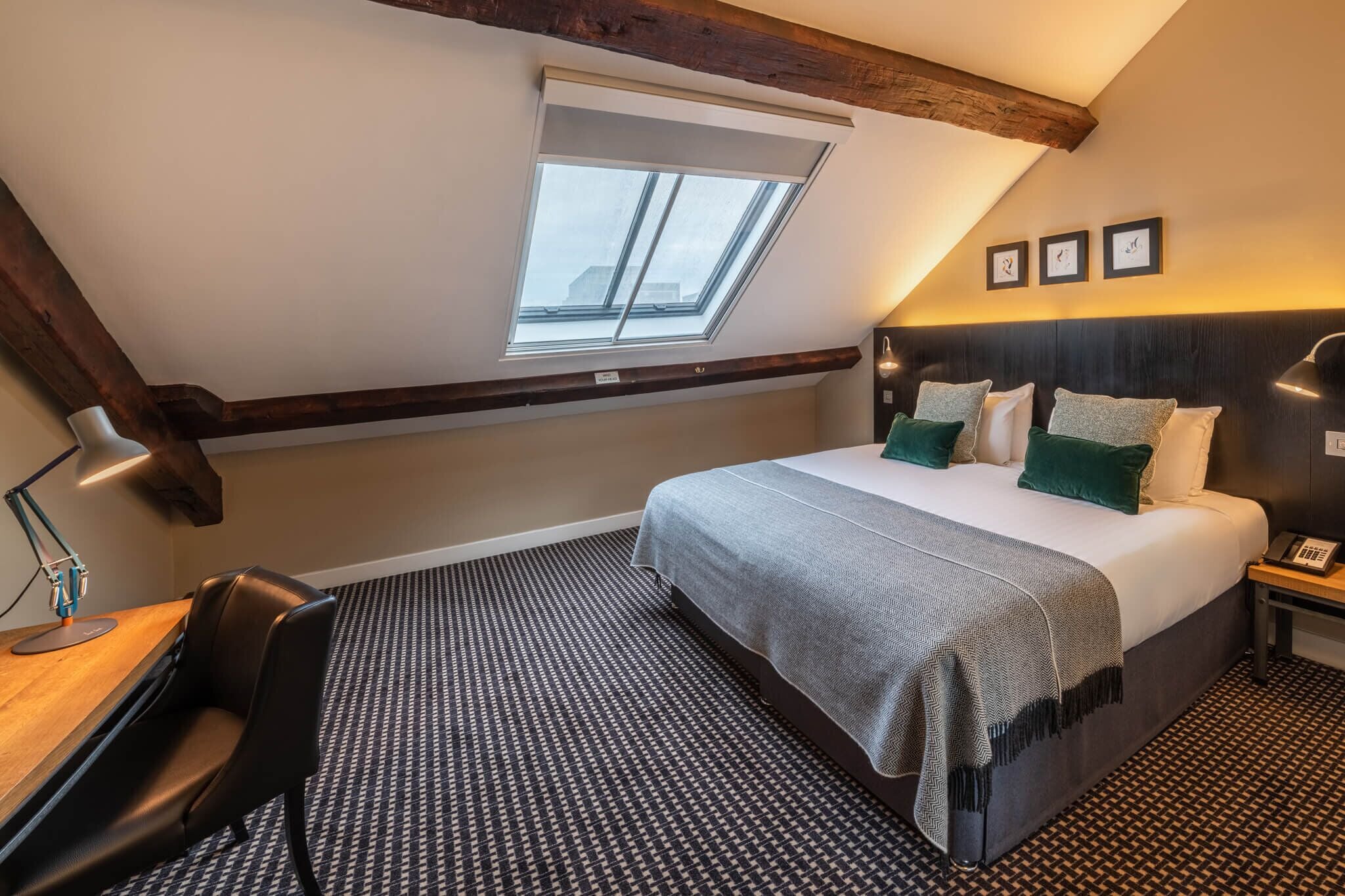 A bedroom with a skylight, a bed with multiple pillows, a desk with a chair, and a patterned carpet.