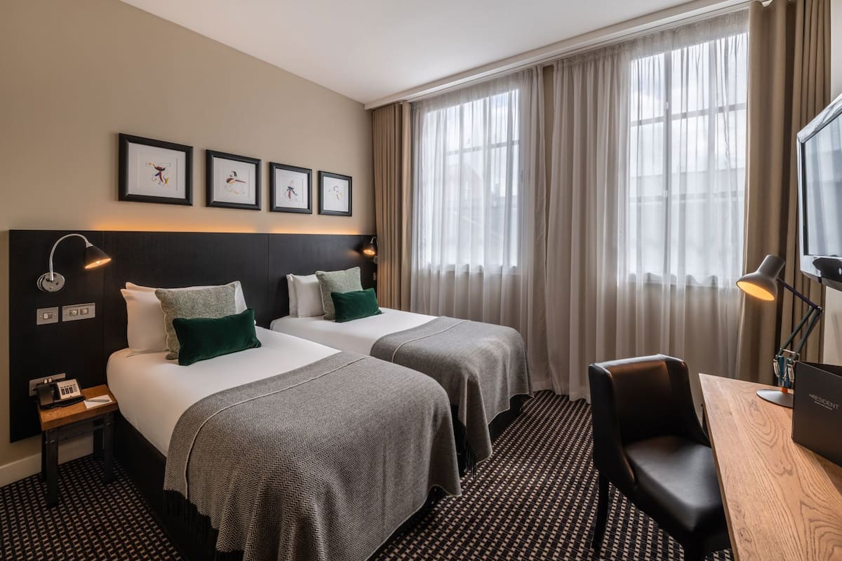 Ett hotellrum med två sängar, ett skrivbord, en stol, en telefon, en lampa och inramade tavlor på väggen.