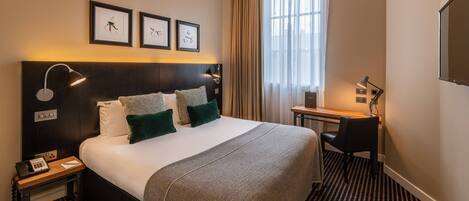 A hotel room with a large bed, a desk, a chair, and a window with curtains.