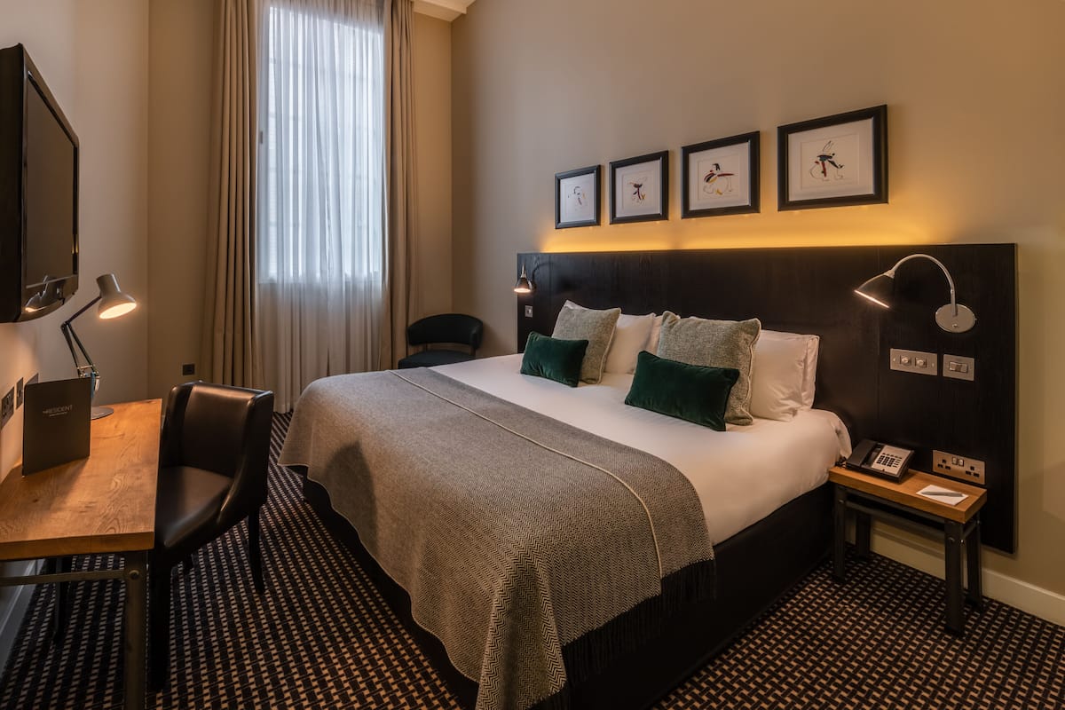 Ett hotellrum med en säng, ett skrivbord, en stol, en tv och ett fönster med gardiner.