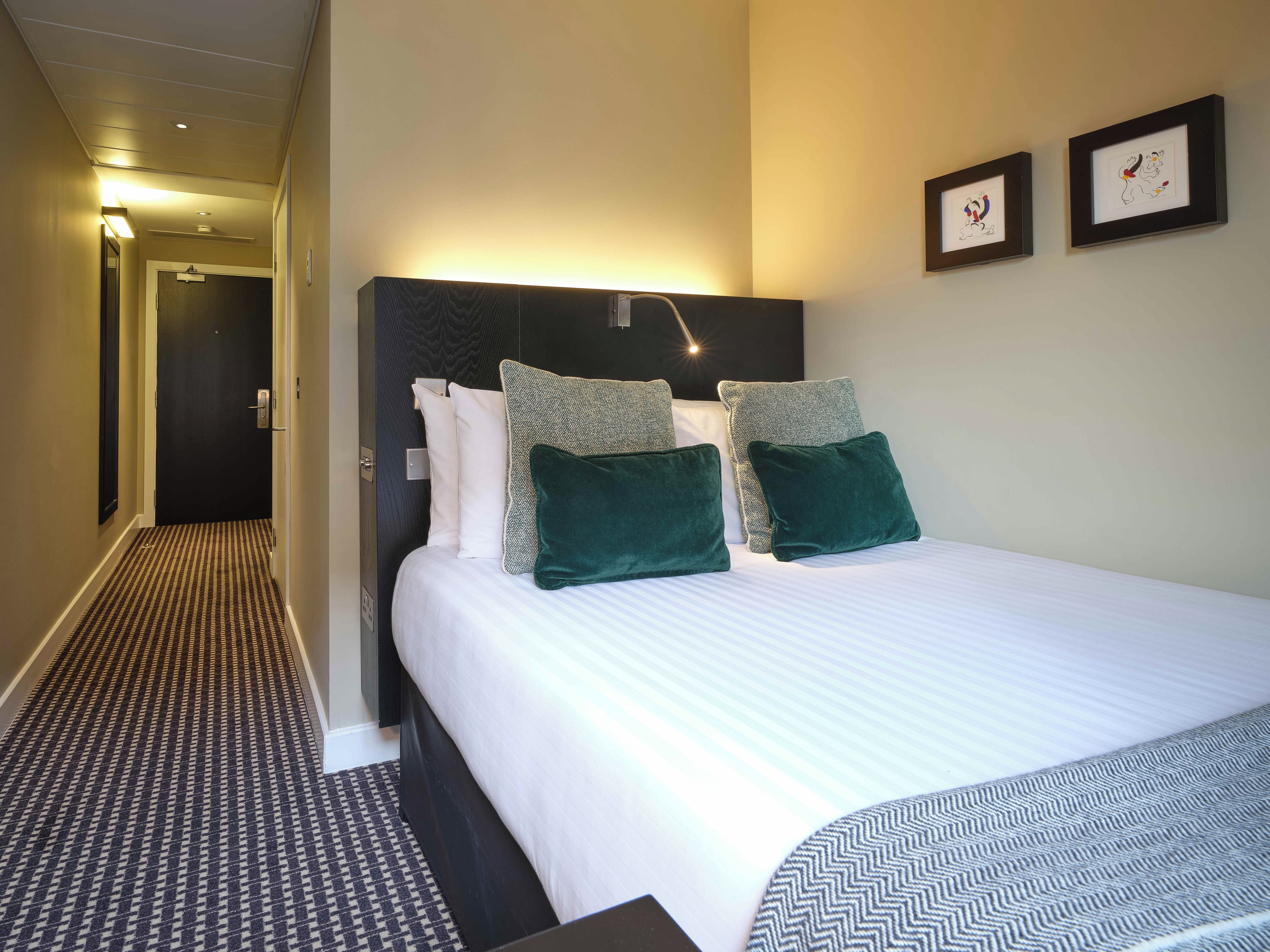 Ein Hotelzimmer mit einem Bett, einem Kopfteil, einem gemusterten Teppich und gerahmten Kunstwerken an der Wand.