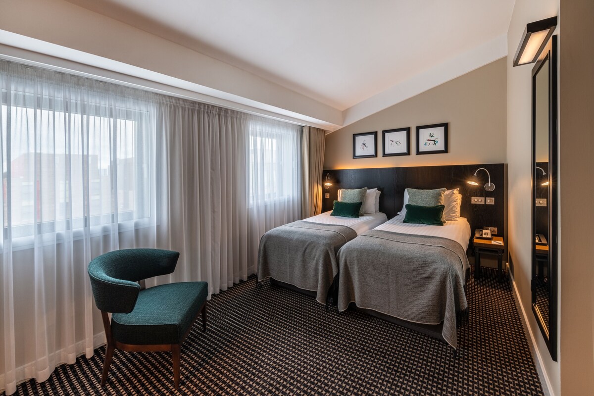 A hotel room with two beds, a chair, a large window with curtains, and framed pictures on the wall.