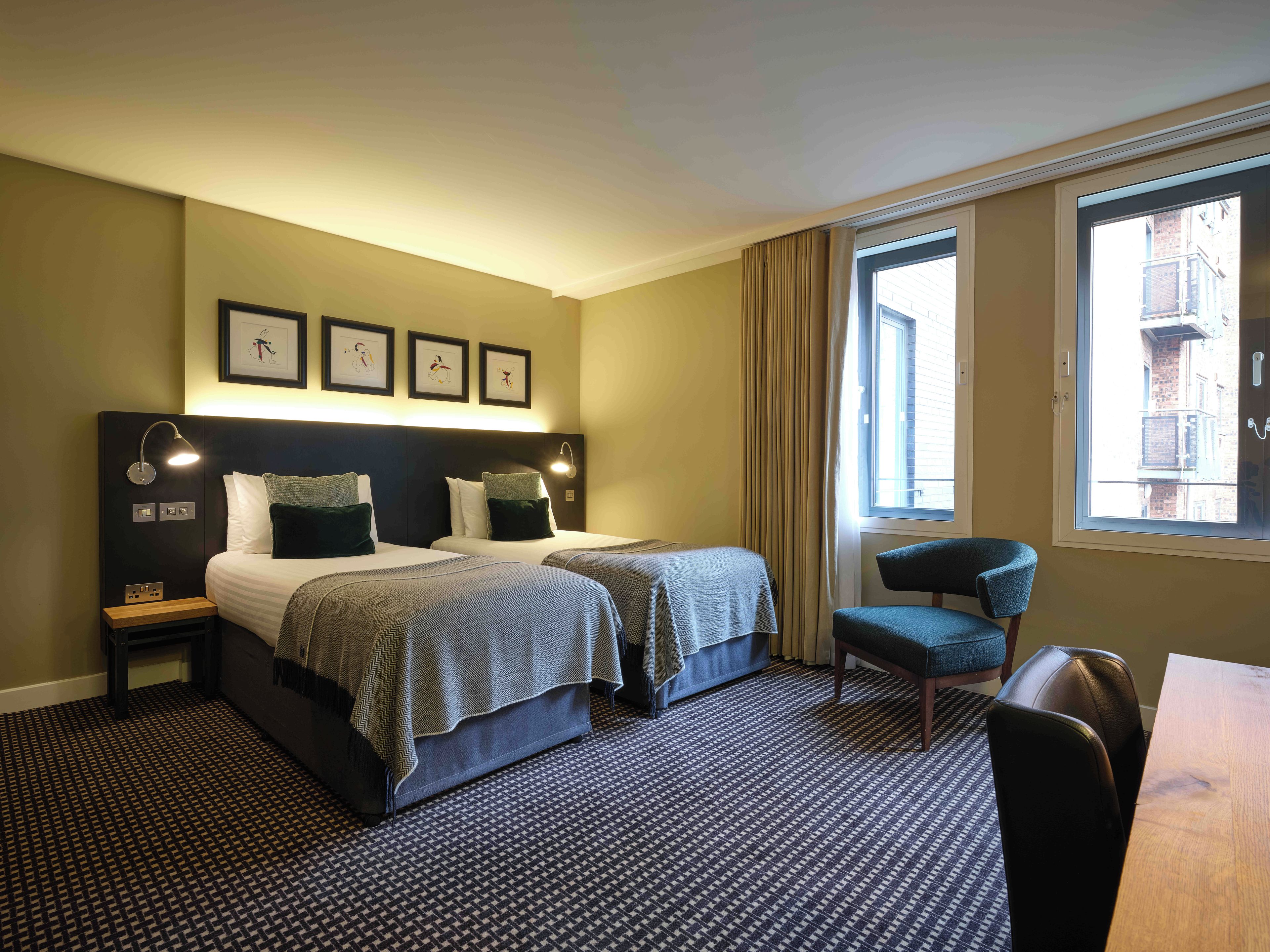 A hotel room with two beds, a desk, a chair, and a window with a view of buildings.