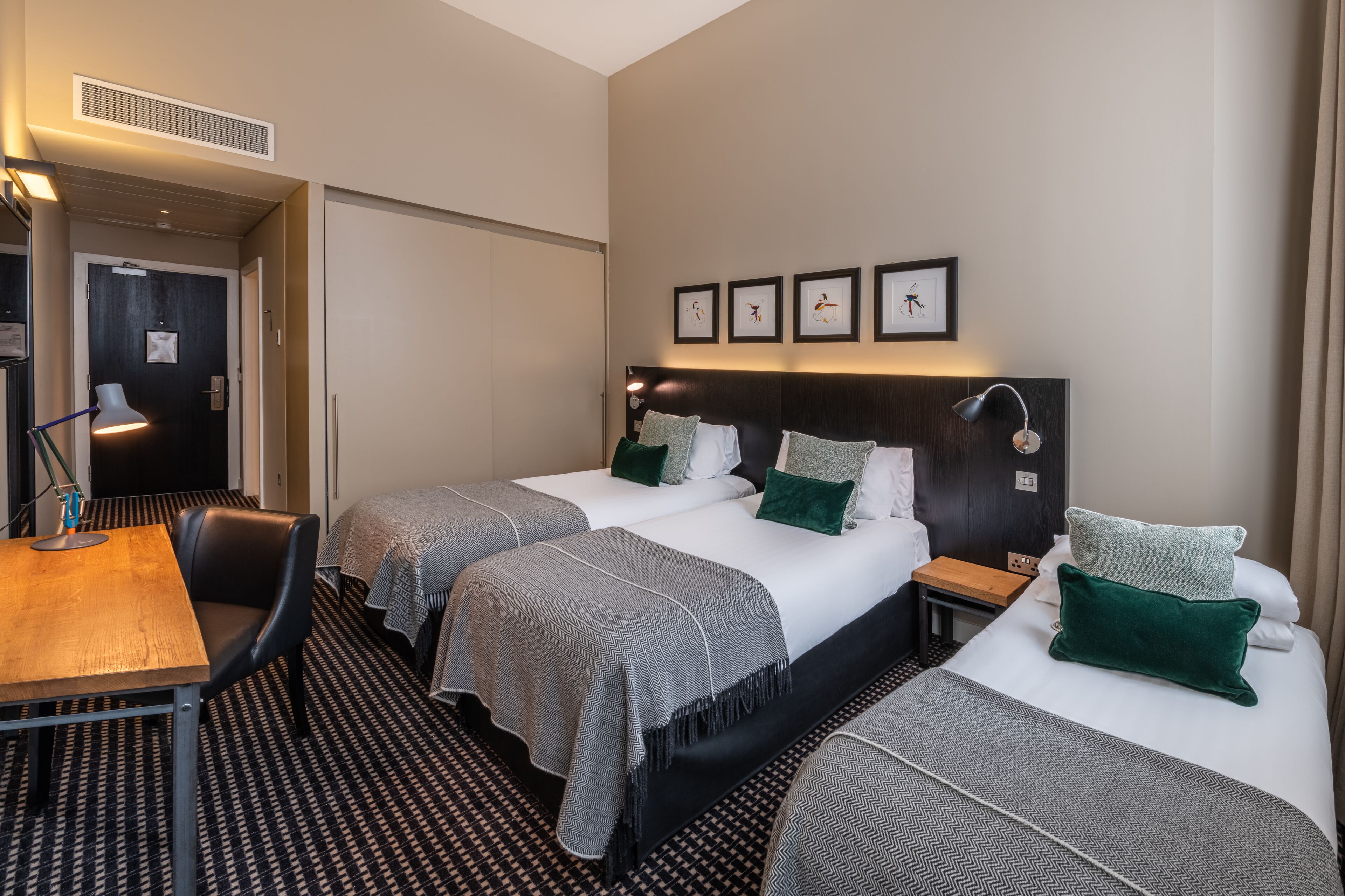 A hotel room with two beds, a desk, and a chair. There are framed pictures on the wall and a lamp on the desk.
