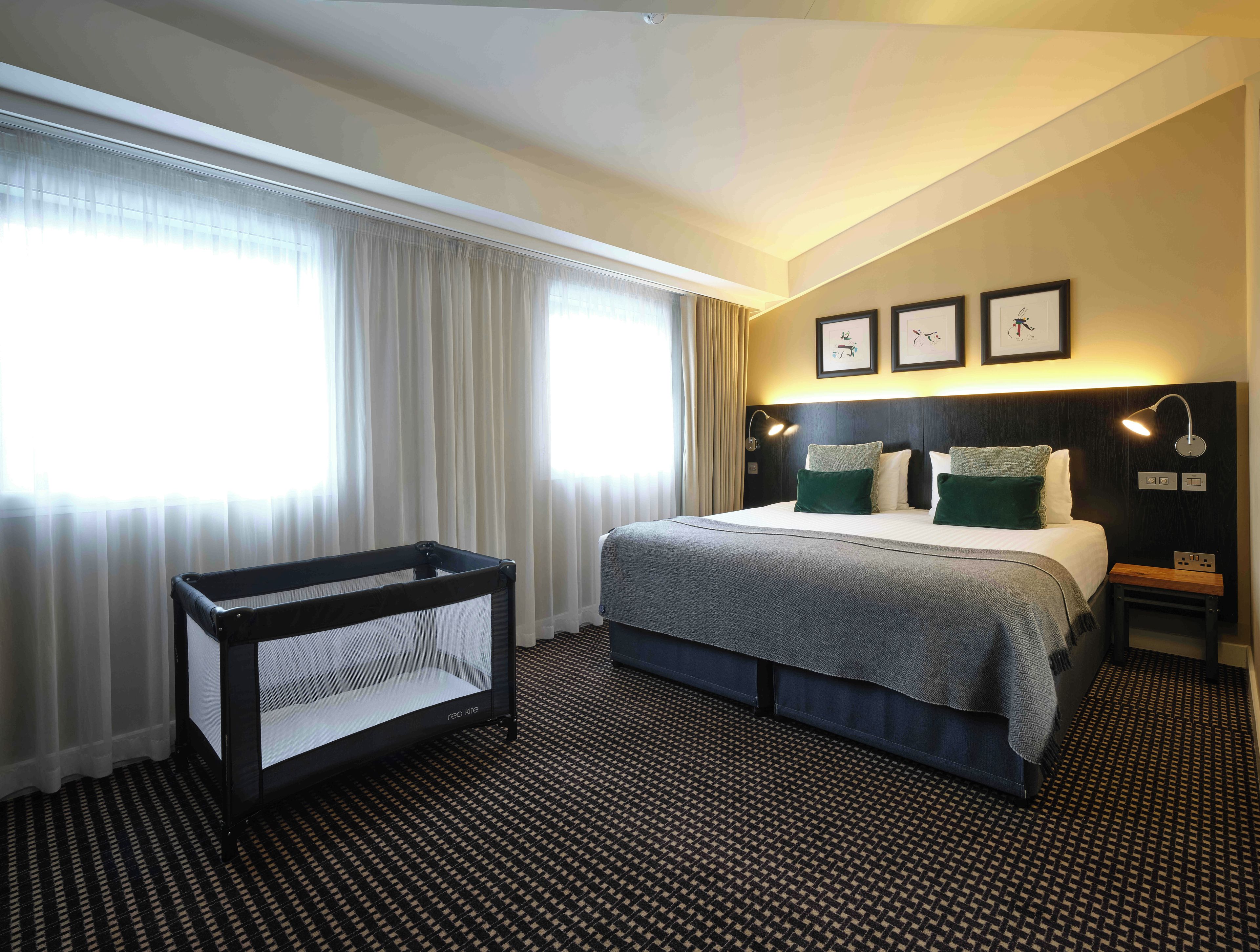 A hotel room with a bed, a crib, a nightstand, and a wall-mounted lamp.