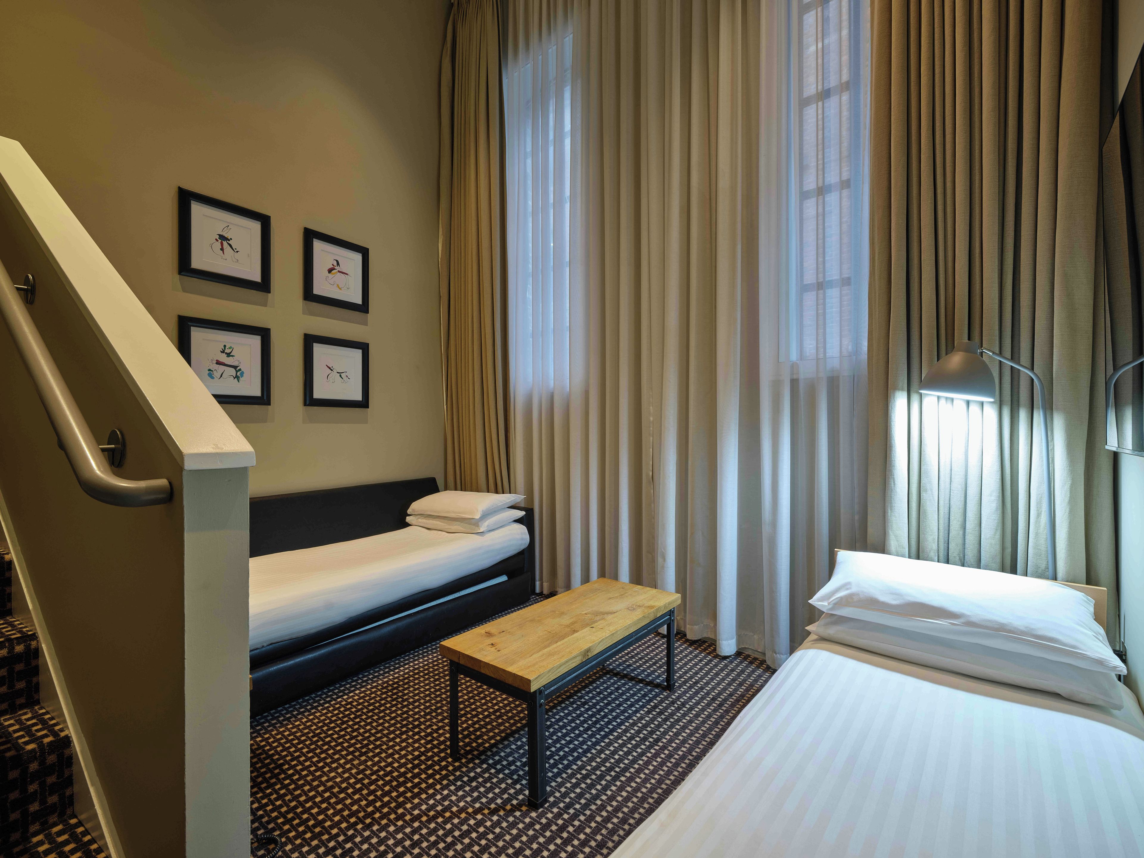 Quarto de hotel com uma cama, um banco, uma escrivaninha e uma escada.