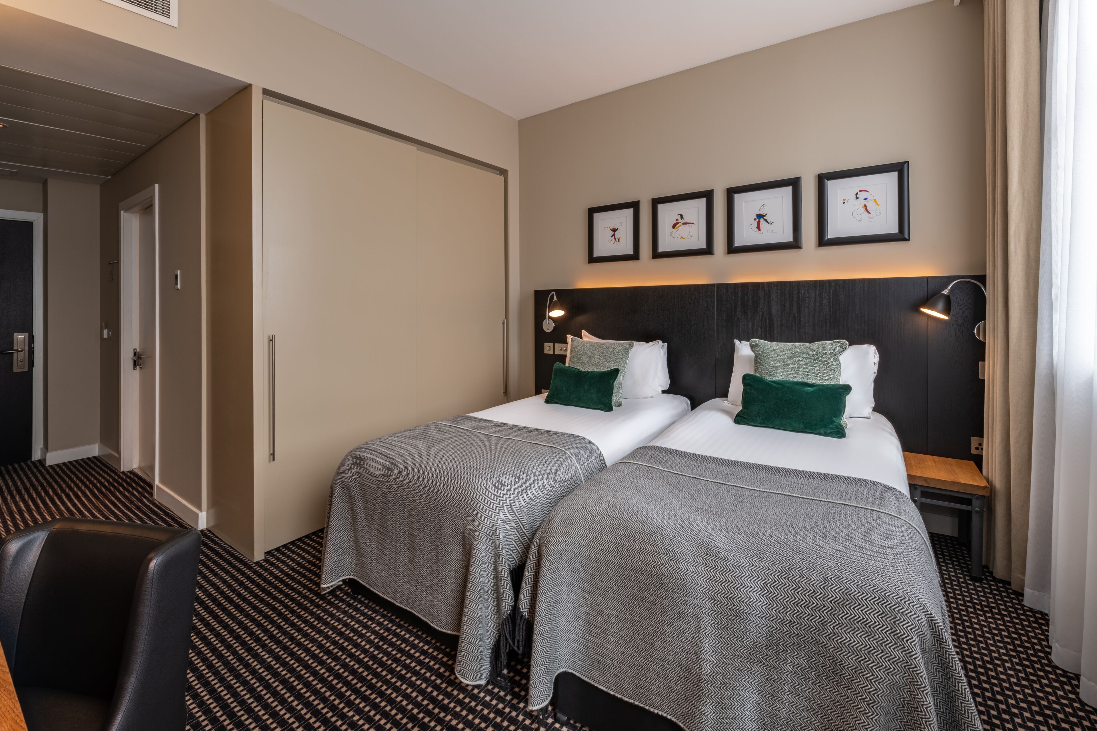 A hotel room with two beds, a desk, and a chair. There are framed pictures on the wall and a door leading to another room.