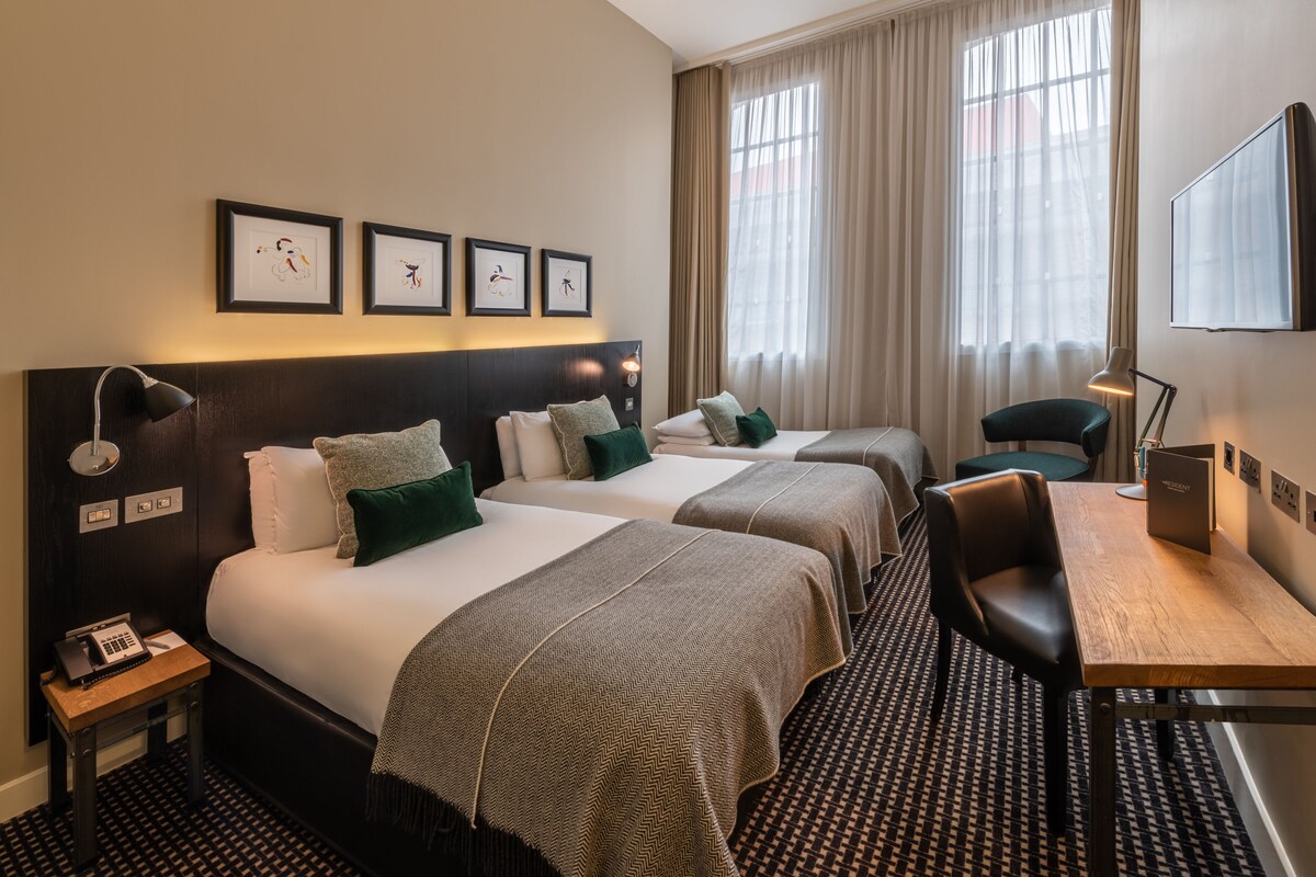 Ein Hotelzimmer mit zwei Betten, einem Schreibtisch, einem Stuhl, einer Lampe und gerahmten Kunstwerken an der Wand.