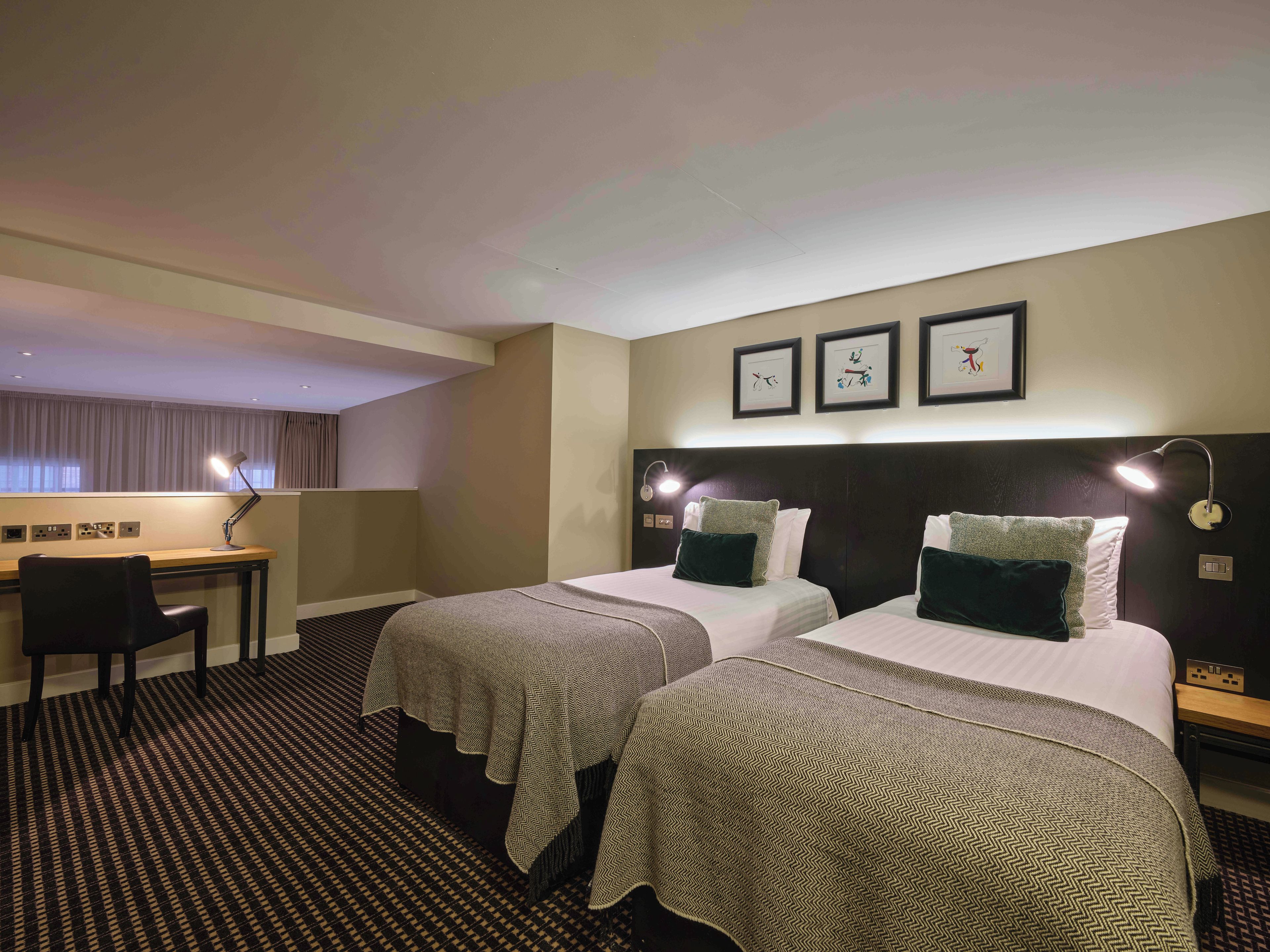A hotel room with two beds, a desk, and a chair. There are wall-mounted lights and a window with curtains.