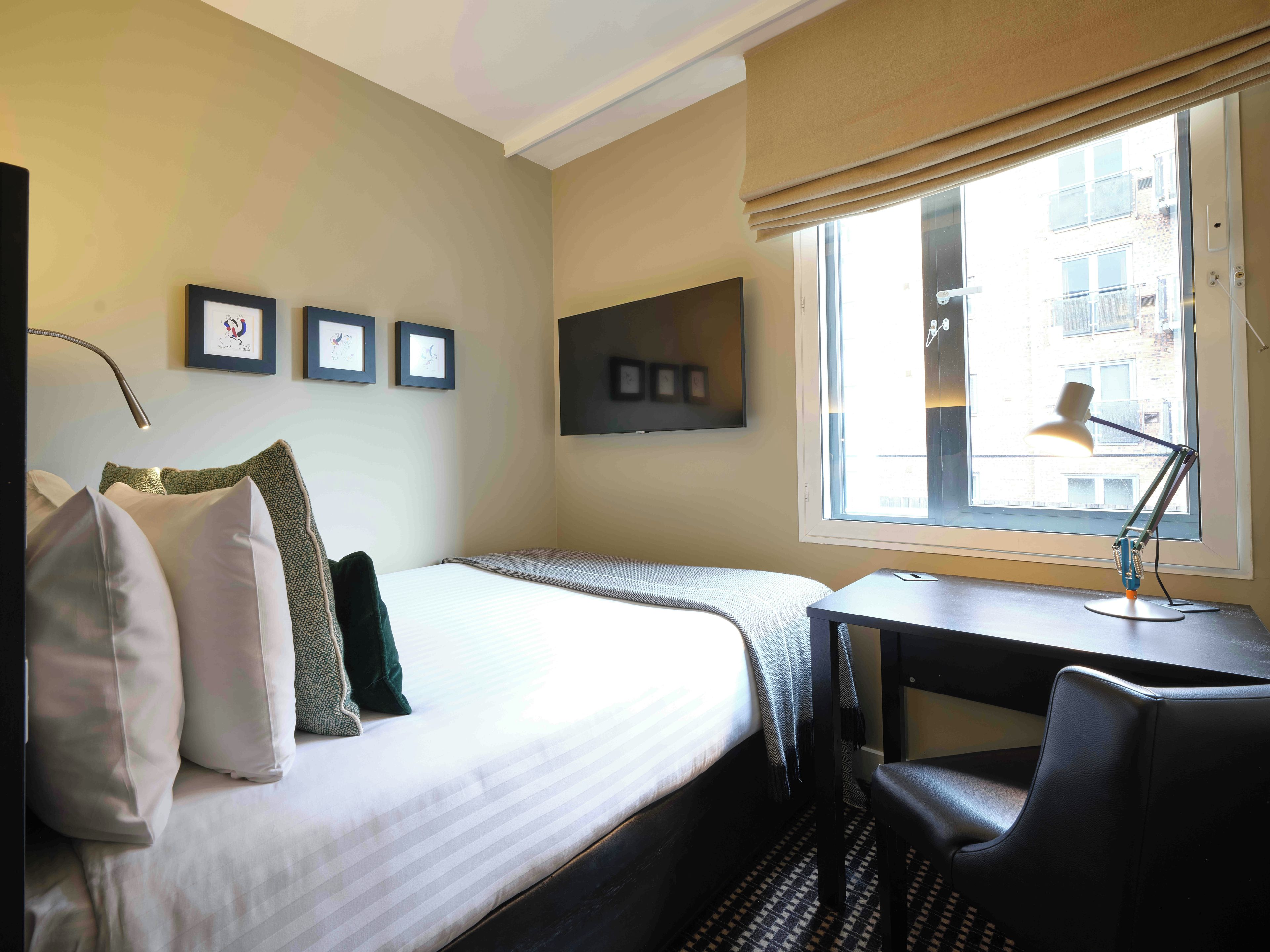 A hotel room with a bed, a desk, a chair, a window with a view of buildings, and framed pictures on the wall.