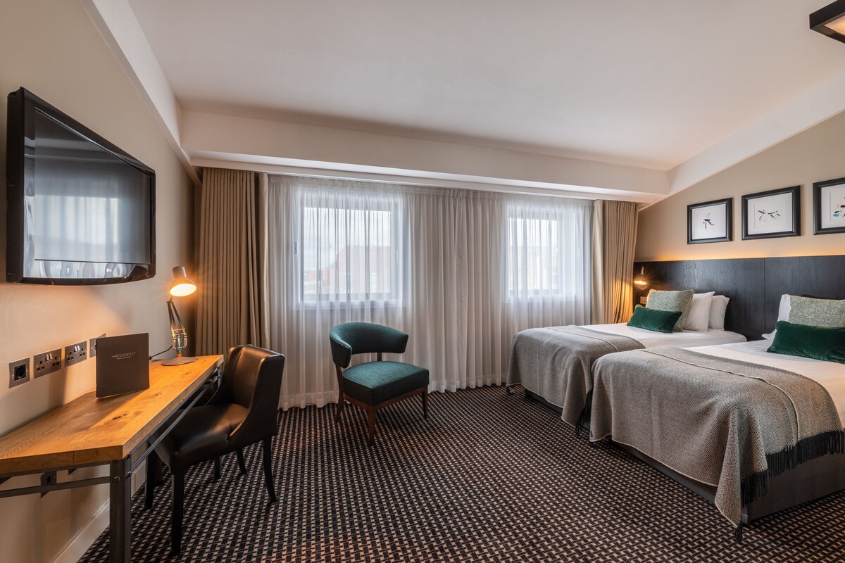 A hotel room with a desk, two beds, a TV, and a view of the outdoors through large windows.