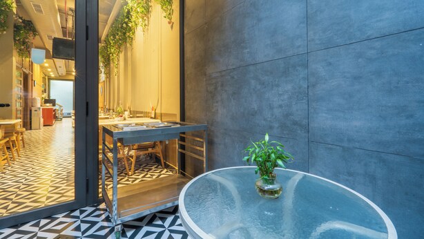 A modern interior with a glass table and a potted plant, a tiled floor, and a view into another room with a kitchen area. - Cosy Grand @Chanakyapuri by The Cosy Hotels (New Delhi)