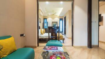 A hotel room with a glass table, a teal sofa, and a bed with a white headboard.