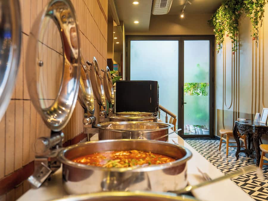 A buffet setup with multiple serving stations and a dining area with a patterned floor and a view of greenery outside.