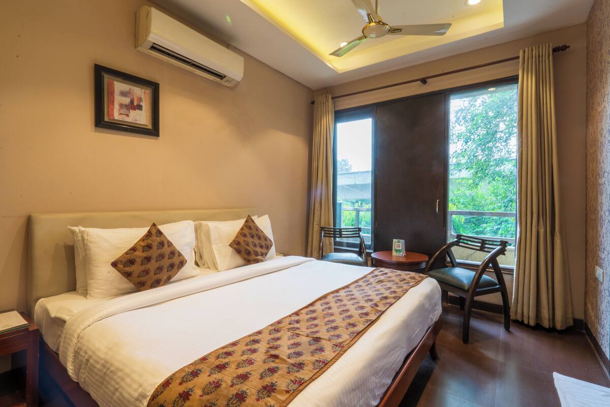 A hotel room with a large bed, a ceiling fan, an air conditioner, a window with curtains, and a chair by the window.
