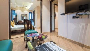 A hotel room with a bed, a desk with a computer, a chair, a table with a bowl of fruit, and a TV mounted on the wall. - Cosy Grand @Chanakyapuri by The Cosy Hotels (New Delhi)