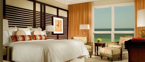 A hotel room with a large bed, a ceiling fan, a view of the ocean, and a window with curtains.