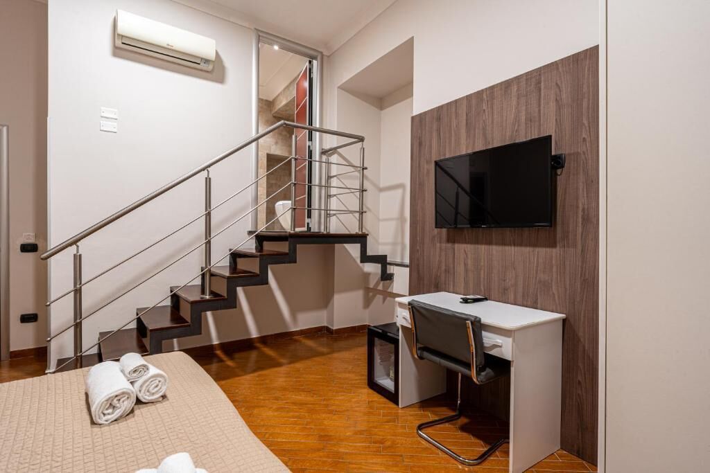 Una habitación de hotel moderna con suelo de madera, un escritorio con una silla, un televisor de pantalla plana montado en la pared, una escalera con barandillas de metal y un par de toallas enrolladas sobre la cama.