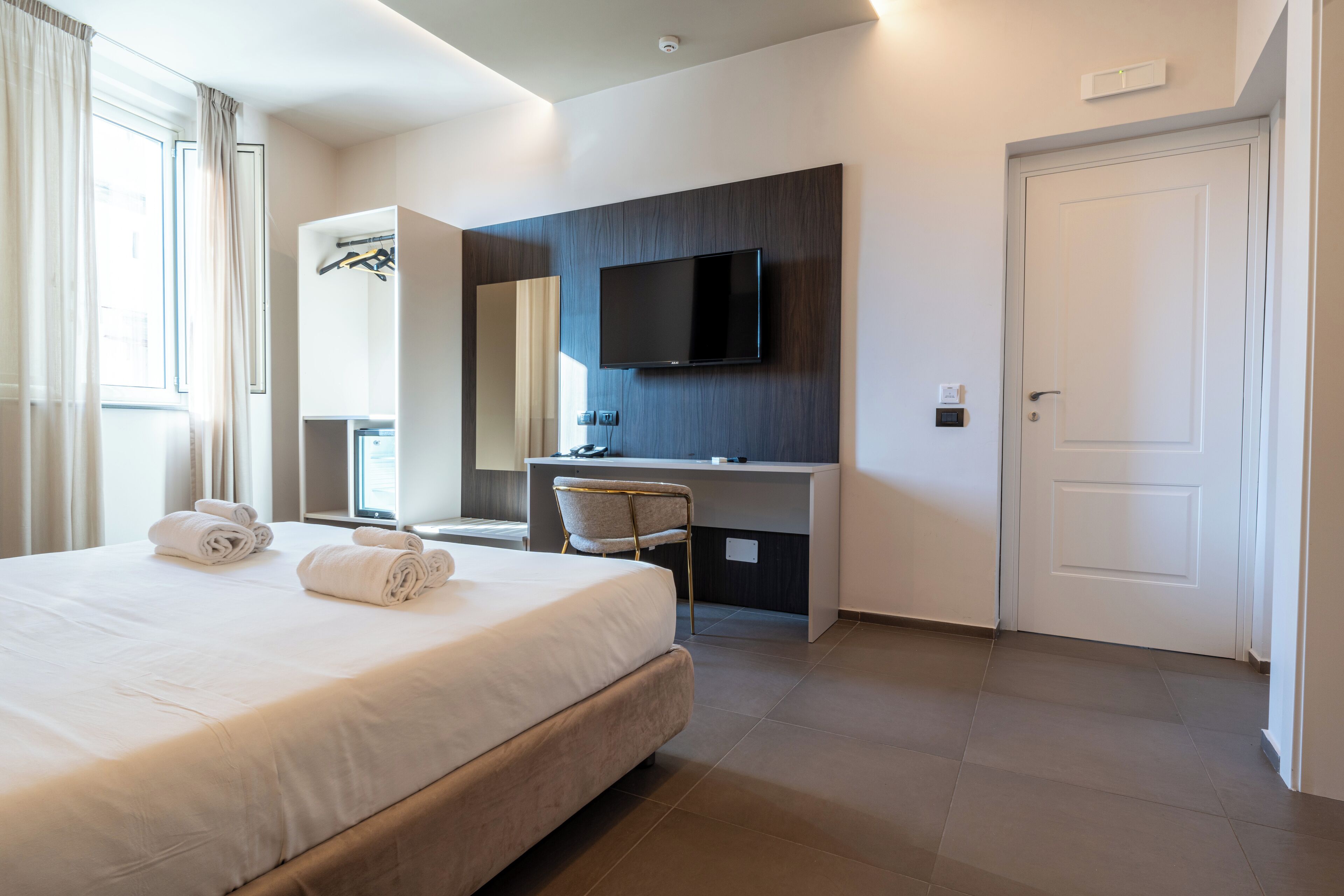 A modern hotel room with a large bed, a desk, a chair, a TV, and a wardrobe.