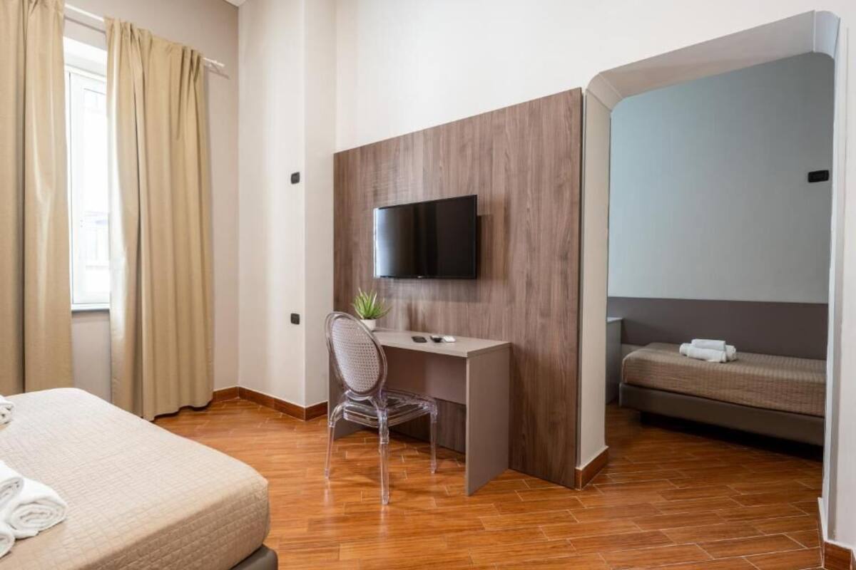 A hotel room with a bed, a desk with a TV, a chair, and a small plant.