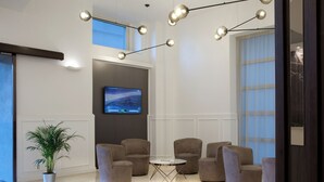 A modern lobby with a round table surrounded by six armchairs, a large flat-screen TV on the wall, and a potted plant.