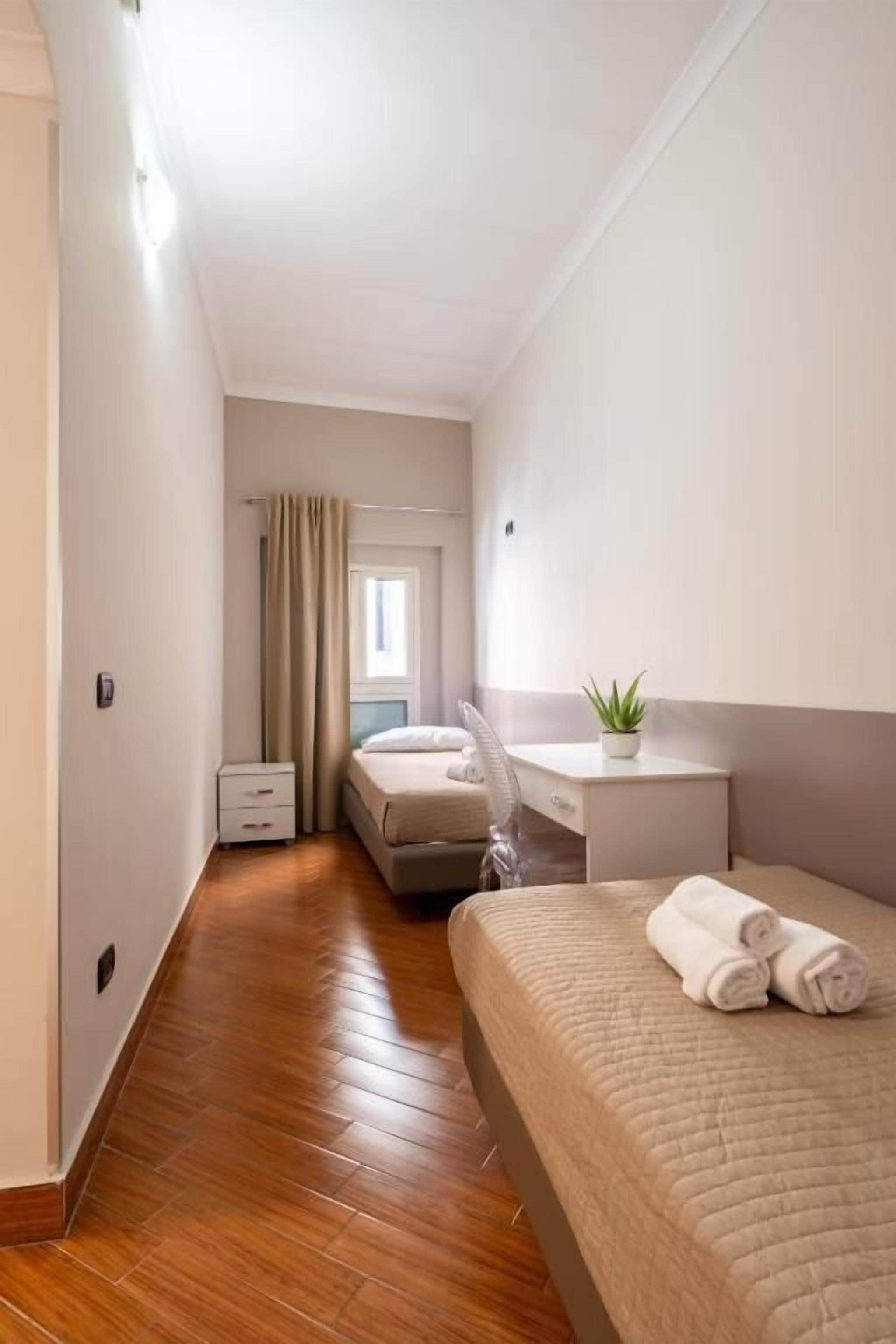 Een smalle hotelkamer met een houten vloer, een bed met een beige sprei, een bureau met een plant en een raam met gordijnen.