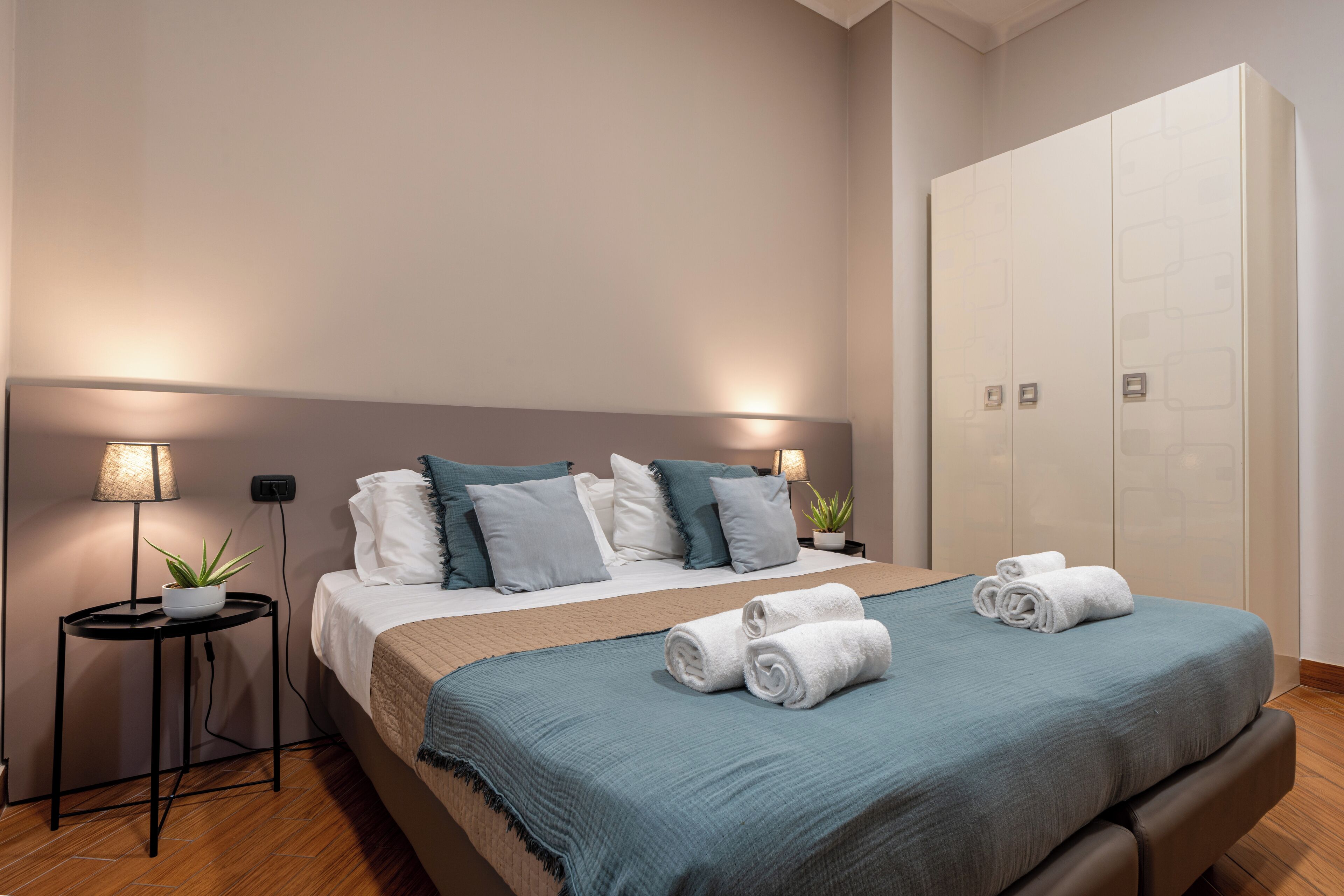 A neatly made bed with a blue blanket, two bedside tables with lamps, and a large wardrobe.
