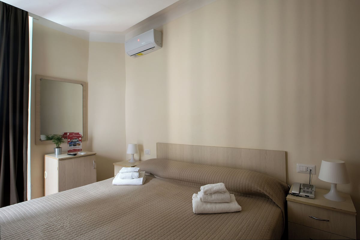 A hotel room with a bed, bedside tables, a desk, a mirror, and an air conditioning unit.