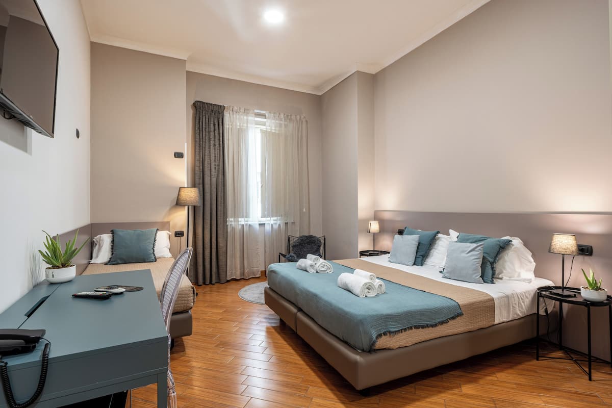 A hotel room with a bed, a desk with a phone, a chair, a small table with a lamp, and a potted plant.