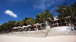 A beachside hotel with stone steps leading to a building, palm trees, and beach chairs with umbrellas. - Novela Muine Resort & Spa (Mui Ne)