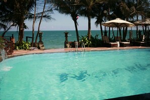 A pool with clear water, surrounded by lounge chairs and umbrellas, with palm trees and a view of the ocean. - Novela Muine Resort & Spa (Mui Ne)