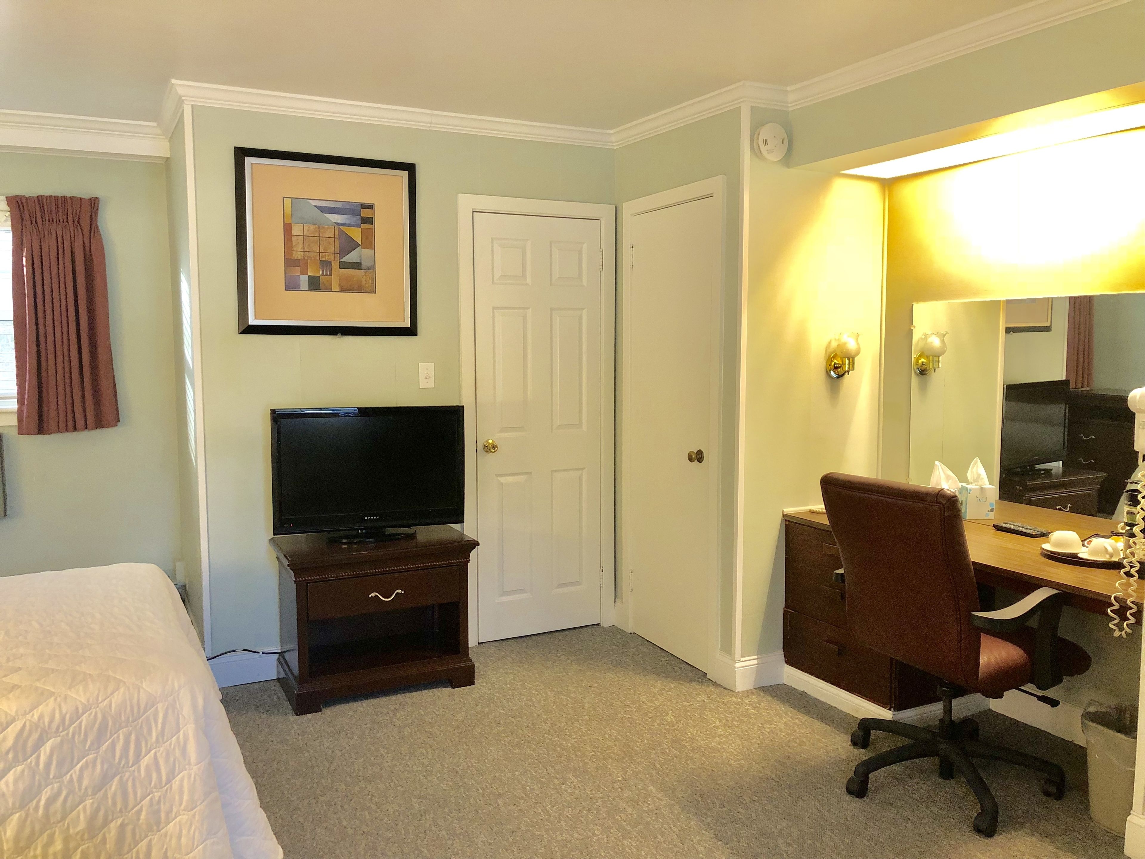 A hotel room with a bed, a TV on a wooden stand, a desk with a chair, a mirror, and a door.