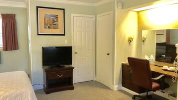A hotel room with a bed, a TV on a wooden stand, a desk with a chair, a mirror, and a door.